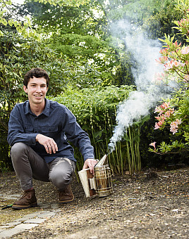 Mateo, in gray pants and a blue collared shirt, crouching on the ground in front of trees and next to a beehive smoker.
