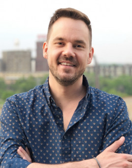 Joshua Ferchau, smiling, arms folded in a blue button-up shirt, stands in front of a city landscape.