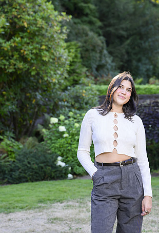 Sonali posing outside in a green garden. She is wearing a white top with buttons and grey pants.