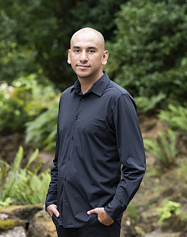 Emmanuel Macias smiling, wearing black shirt, forest background.