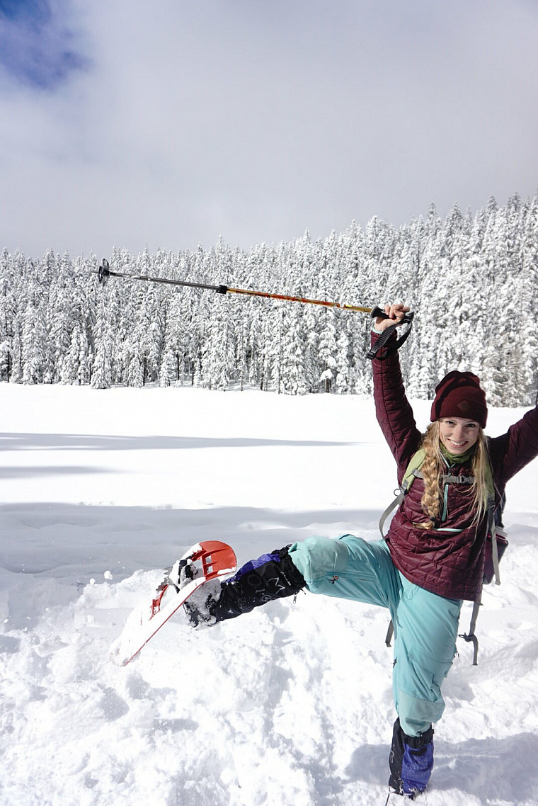 Angelina puts her leg in the air while snowshoeing with snow covered trees behind her.