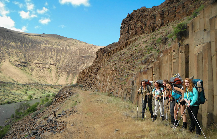 Image shows people wearing backpacks on a trail with columnar basalt cliffs behind them and the Deschutes river below