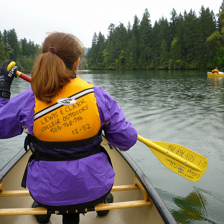 A student in a purple jacket and yellow PFD canoes on a lake with trees in the distance