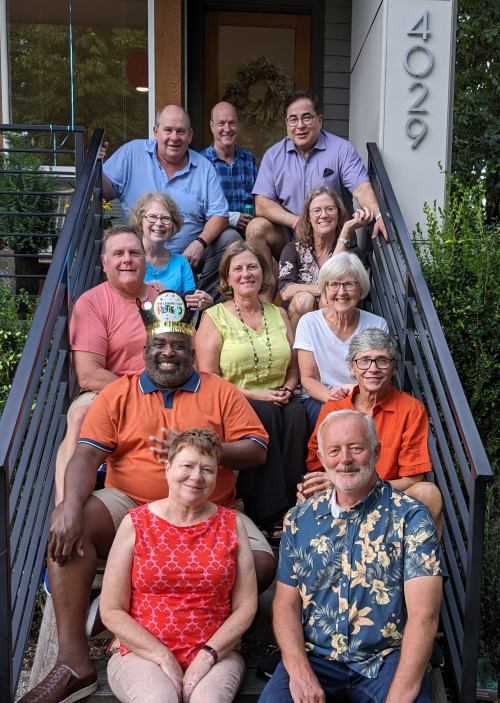 Several smiling adults seated on outdoor stairs leading to the front door of a residence. Older people dressed for warm weather.