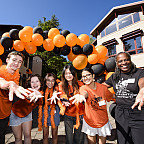 The president and a group of students smiling and reaching their hands out to the camera. A black and orange balloon arch is behind them.