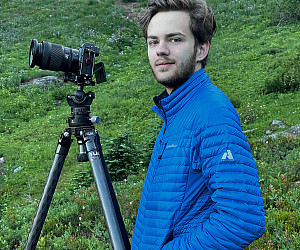 Nicholas standing outside in a blue jacket, next to a camera on a tripod.