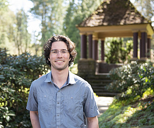 Cooper posing outside near a gazebo, wearing a blue button-up short sleeve shirt and glasses.