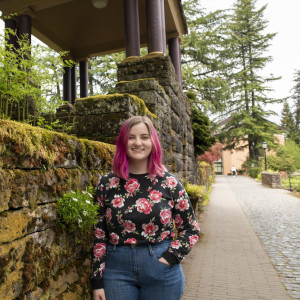 Sophia Reeves BA '23 poses outside. She has long pink hair and is wearing a black shirt with pink flowers and blue jeans.