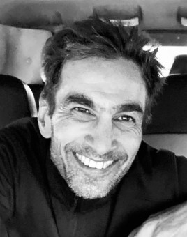 Kabir is smiling at the camera with his arms crossed in a black and white photo